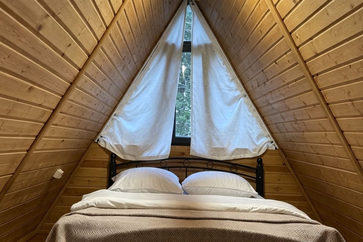 Techo de madera triangular con una cama en el centro, estructura metálica y ropa de cama blanca.
