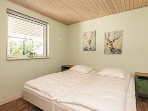 Un dormitorio con una cama, una ventana con persianas, una mesita pequeña y dos cuadros colgados en la pared.