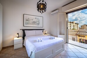 A modern bedroom with a large bed, a nightstand, a bedside lamp, a framed picture on the wall, and a view of the city through a balcony. - Garella (Prato)