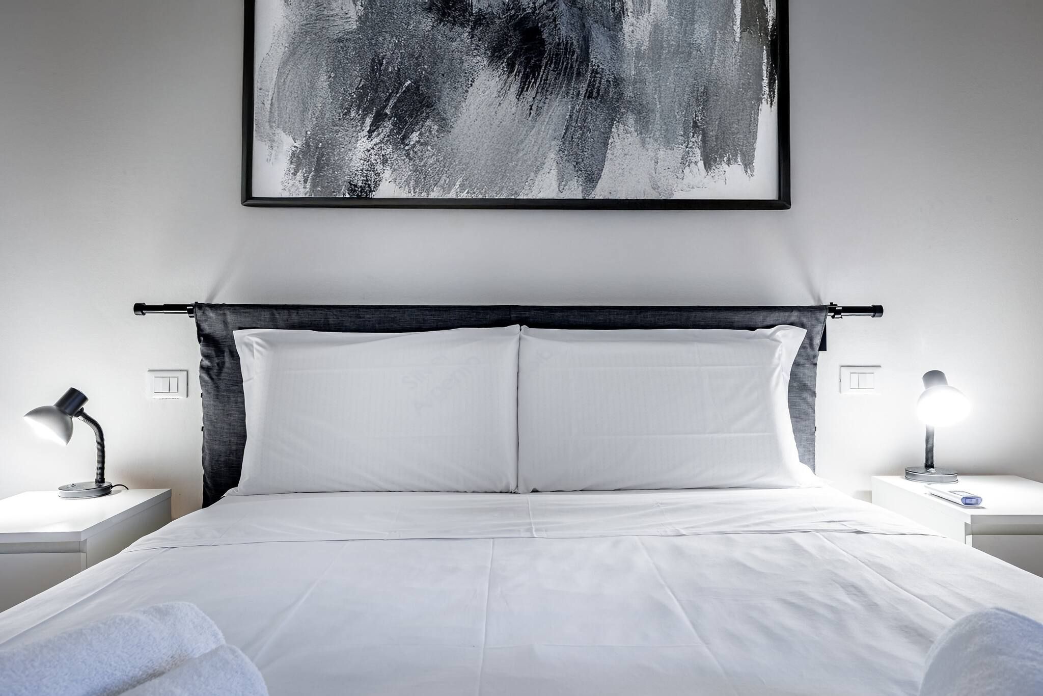 A modern hotel room with a large bed, two bedside tables, and a black abstract painting on the wall.