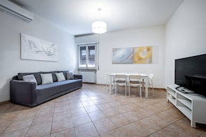 Un saló modern amb un sofà gris, taula de dinar blanca i cadirees, televisió de pantalla plana i obra d'art abstracta a la paret.