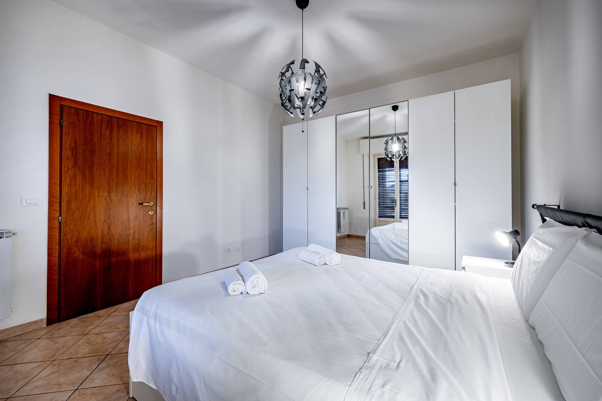 A hotel room with a wooden door, a large bed with white linens, a mirror, and a chandelier.