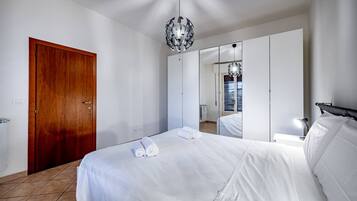 A hotel room with a wooden door, a large bed with white linens, a mirror, and a chandelier.