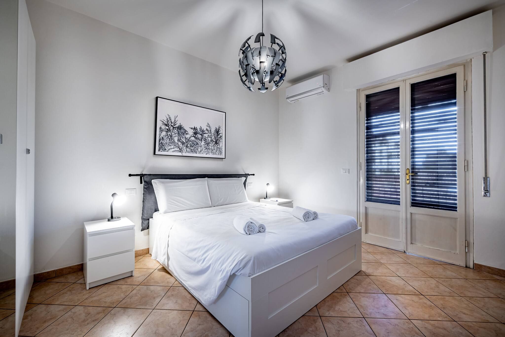 Un dormitorio con cama, mesitas de noche, lámpara colgante, ventana con persianas y aire acondicionado instalado en la pared.