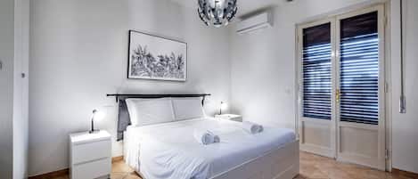 A bedroom with a bed, bedside tables, a hanging light fixture, a window with blinds, and a wall-mounted air conditioner.