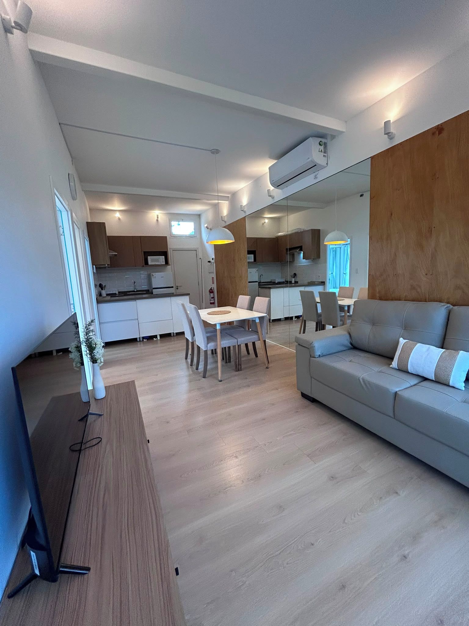 Una sala de estar moderna con sofá y comedor, suelo de madera y una cocina al fondo.