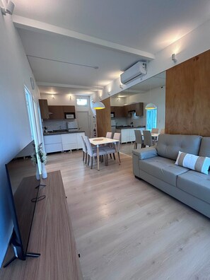 Sala de estar moderna com sofá e área de jantar, piso de madeira e cozinha ao fundo.