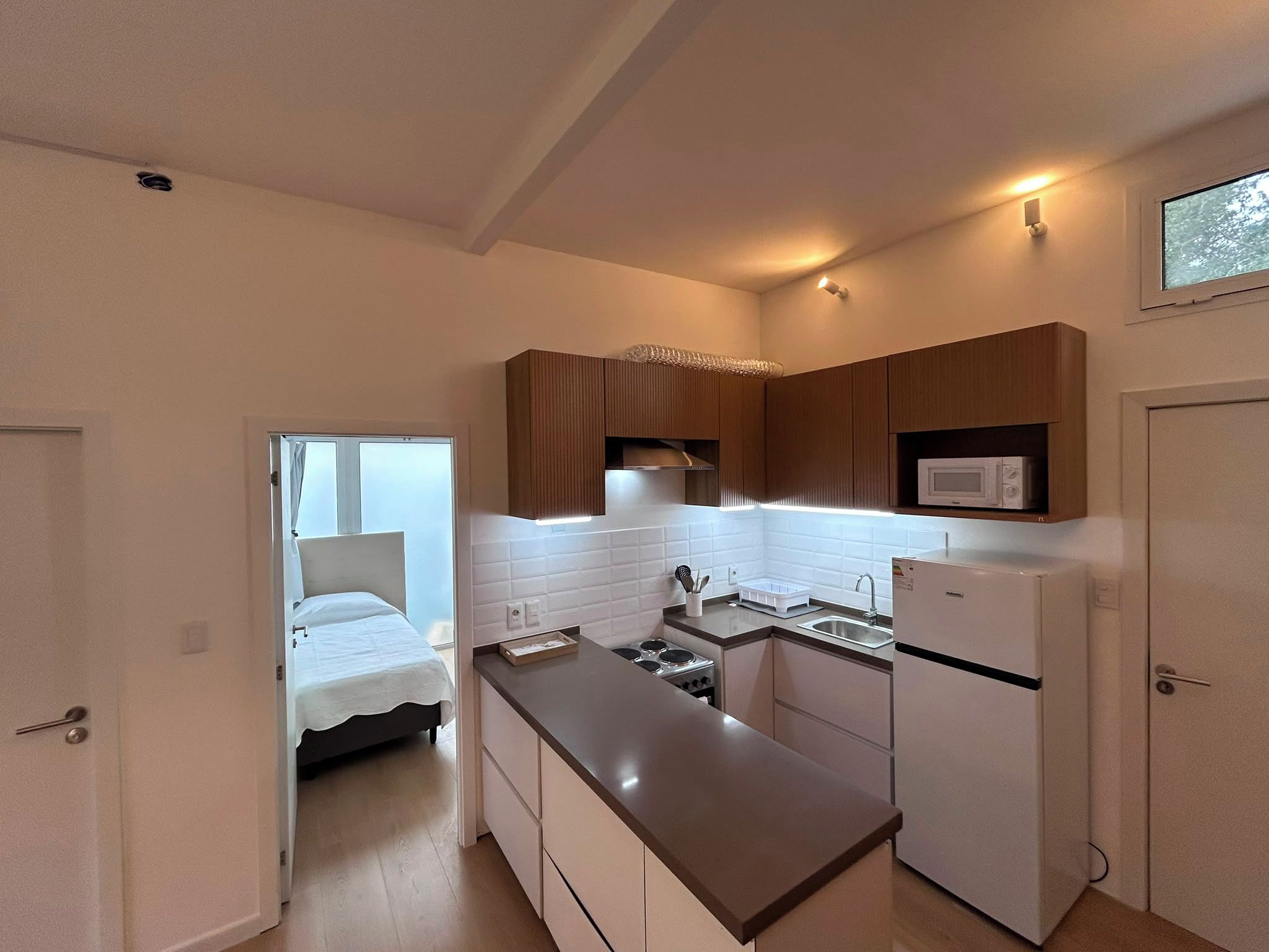 Cocina compacta con estufa, microondas y refrigerador pequeño. Se ve una cama a través de una puerta abierta, lo que sugiere un kitchenette o alojamiento tipo dormitorio.