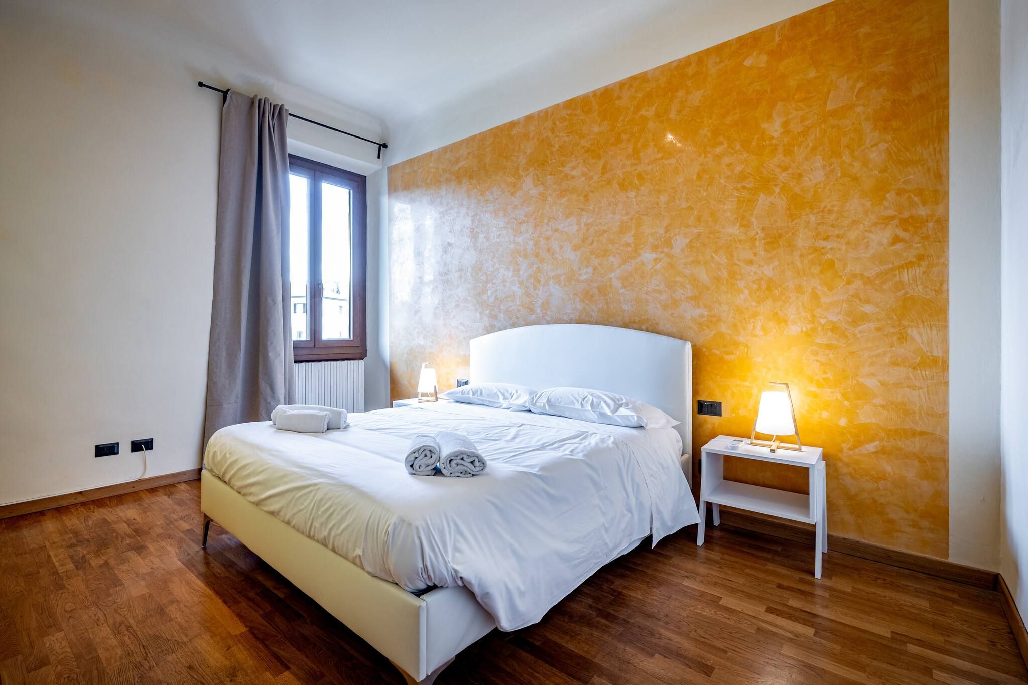 Un dormitorio con cama, mesita de noche, lámpara y ventana con cortinas.