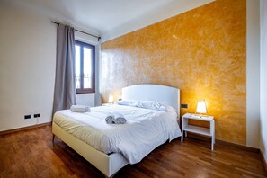 Quarto com cama, mesa de cabeceira, luminária e janela com cortinas.