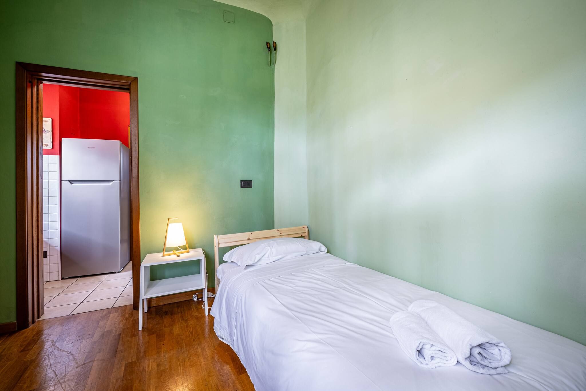 Una habitación pequeña con cama individual, piso de madera, mesita de noche con lámpara y una puerta con marco rojo que da a una zona de cocina.