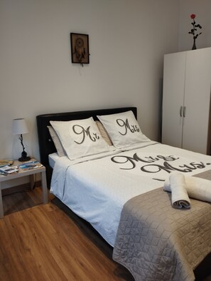 Ein ordentlich bezogenes Bett mit einer gesteppten Decke und Kissen mit der Aufschrift 'Herr' und 'Frau'. Ein Nachttisch mit Lampe und Büchern ist zu sehen. Im Hintergrund steht ein Kleiderschrank.
