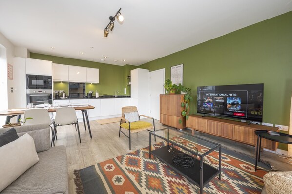A modern living room with a flat-screen TV, a dining area with a kitchen in the background, and a green accent wall. - The Greenwich Green | 2BR • 2BA • Balcony - Pass the Keys (London)