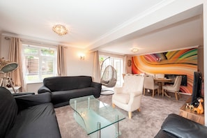 A modern living room with a glass coffee table, a black sofa, a white armchair, and a colorful abstract wall mural. - The Willow House | 4BR • Garden • Parking for 2 - Pass the Keys (Sidcup)