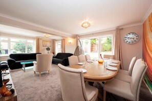 A well-lit living room with a dining area, featuring a large window, a chandelier, and a clock on the wall. - The Willow House | 4BR • Garden • Parking for 2 - Pass the Keys (Sidcup)