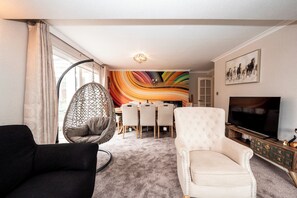 Una sala de estar con un sofá negro, un sillón blanco, una silla colgante, un comedor con un mural colorido y un televisor sobre un mueble de madera.