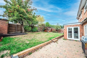 A backyard with a grassy lawn, a wooden fence, and a brick patio. - The Willow House | 4BR • Garden • Parking for 2 - Pass the Keys (Sidcup)