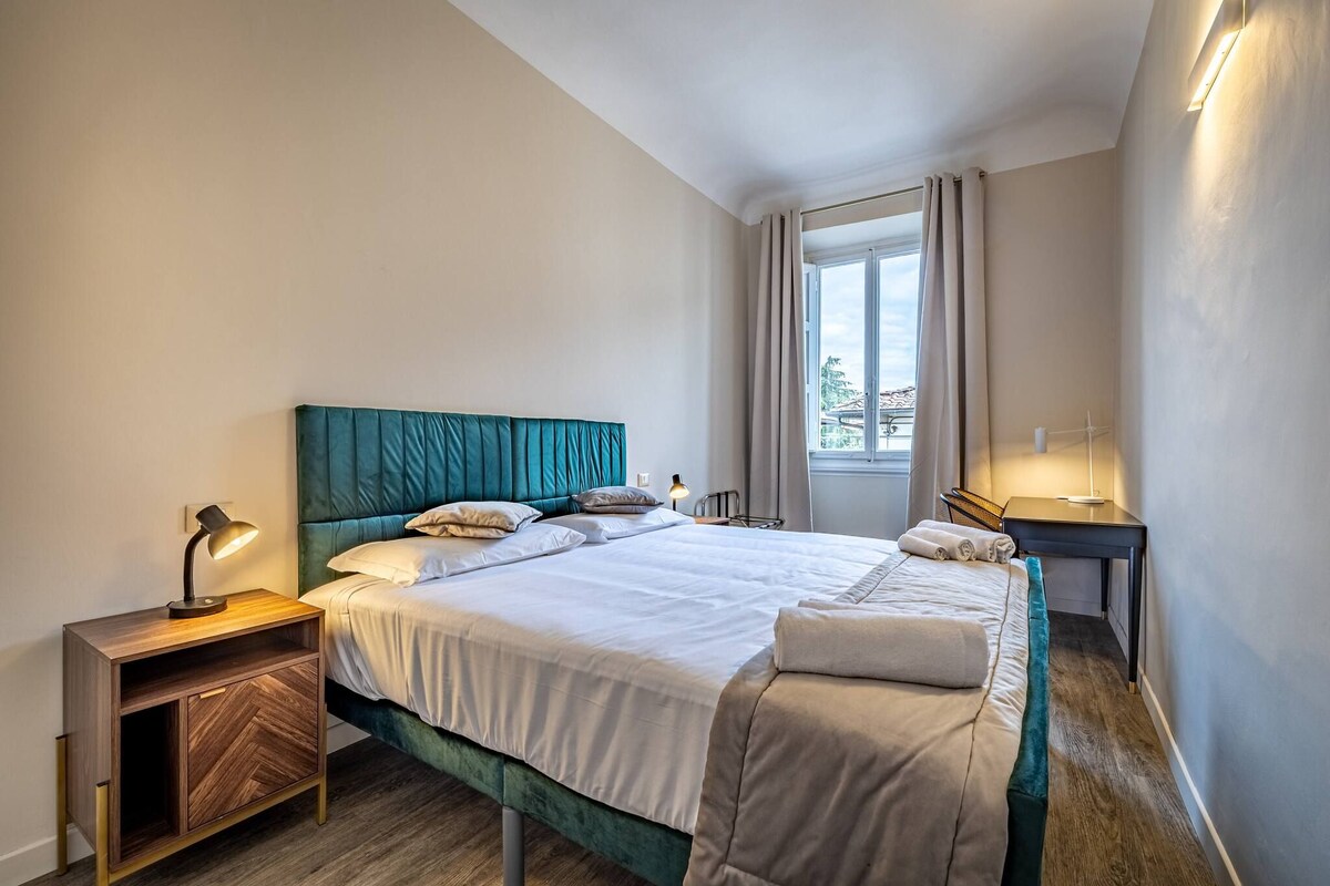 A neatly made bed with a teal headboard, a bedside table with a lamp, and a window with curtains.