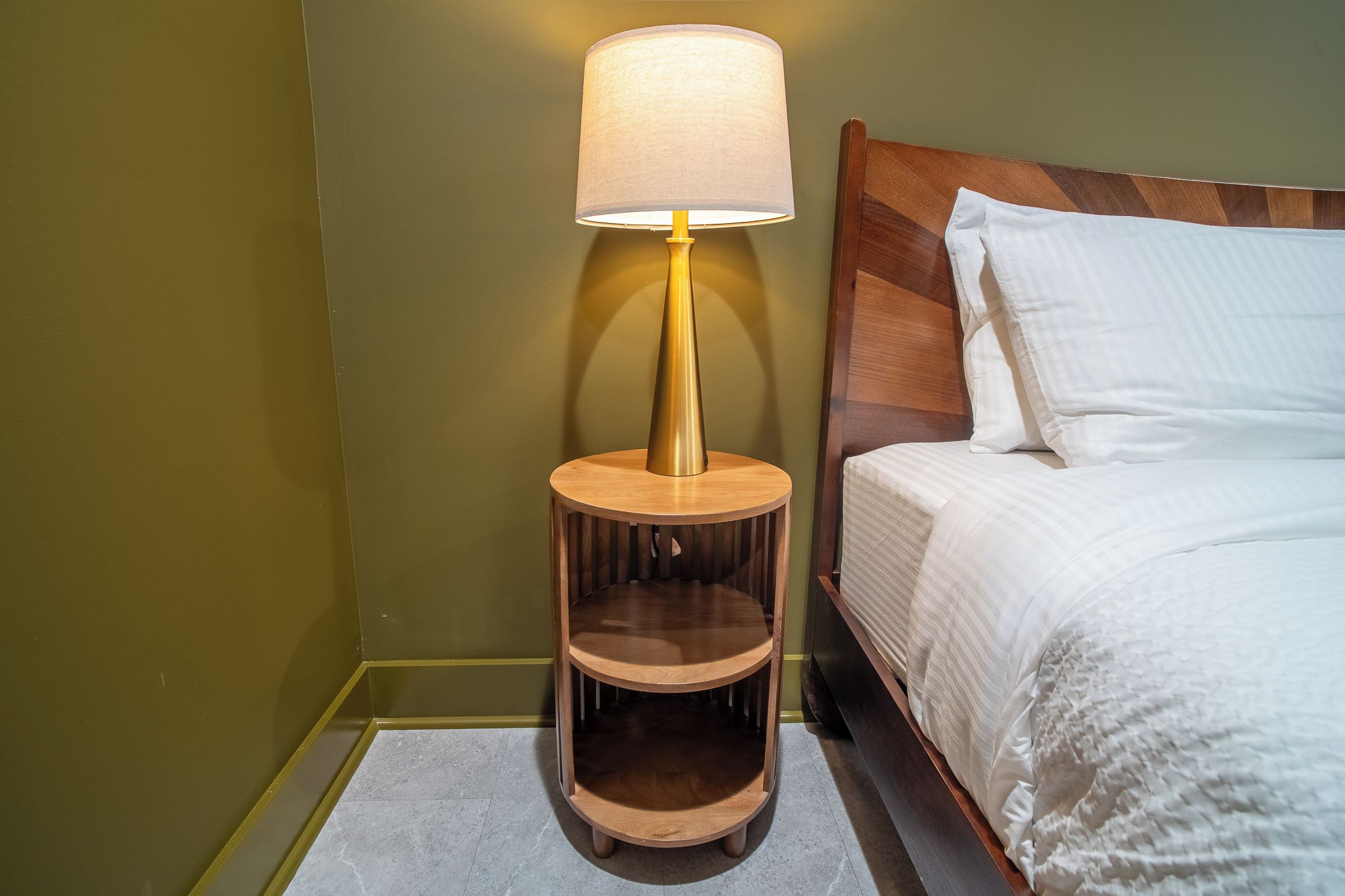 Un lit avec une literie blanche, une table de chevet en bois avec une lampe et une petite étagère en bois.
