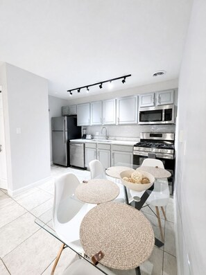 A modern kitchen with white cabinets, stainless steel appliances, and a glass dining table with woven placemats. - Across from Shands | 2BR Condo Near UF + Pool (Gainesville)