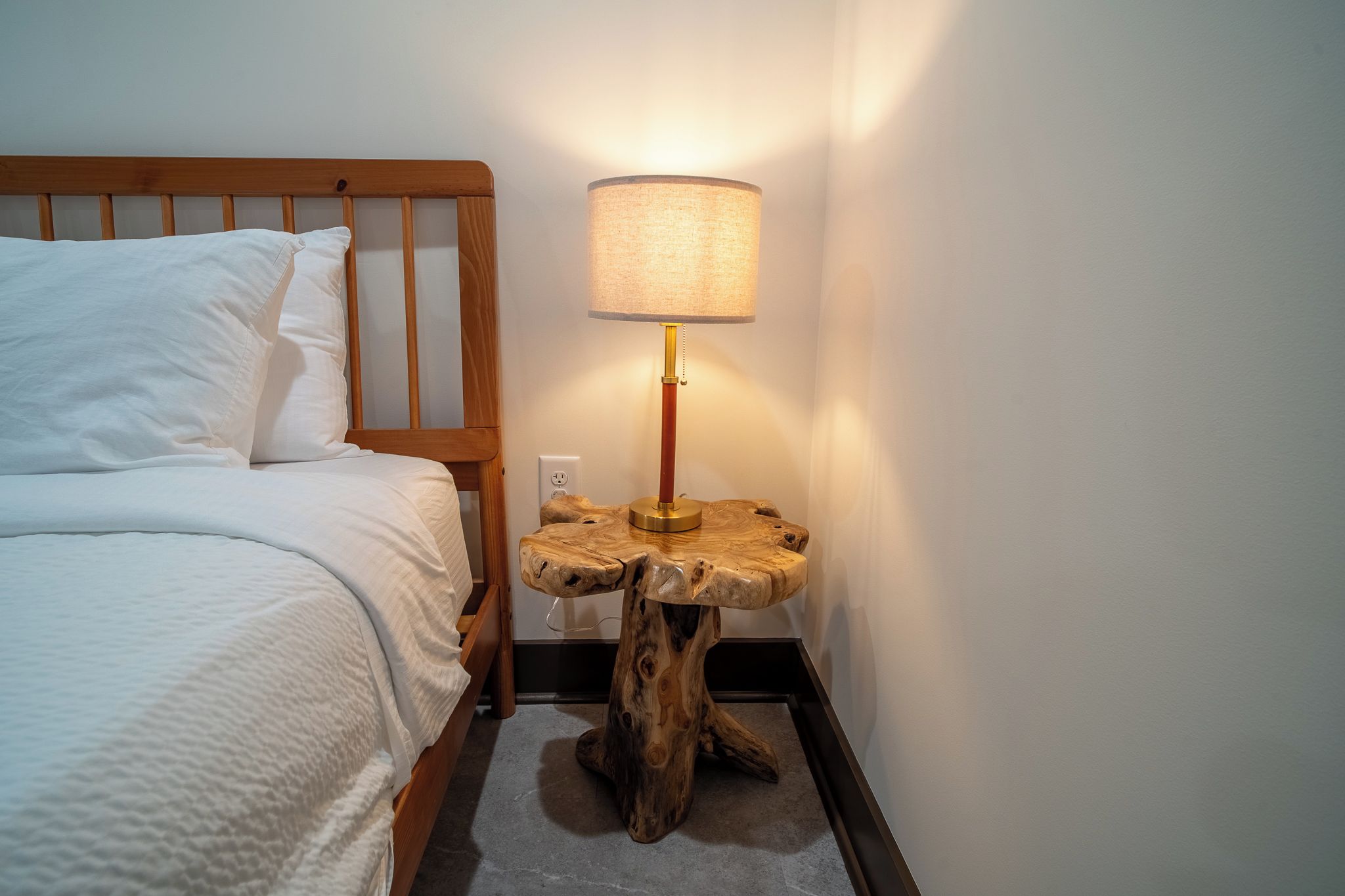 Ein ordentlich bezogenes Bett mit weißen Leinen und Kissen, ein Nachttisch aus Holz mit einer Lampe und eine Steckdose.