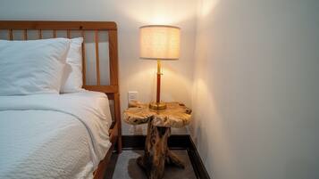 Ein ordentlich bezogenes Bett mit weißen Leinen und Kissen, ein Nachttisch aus Holz mit einer Lampe und eine Steckdose.