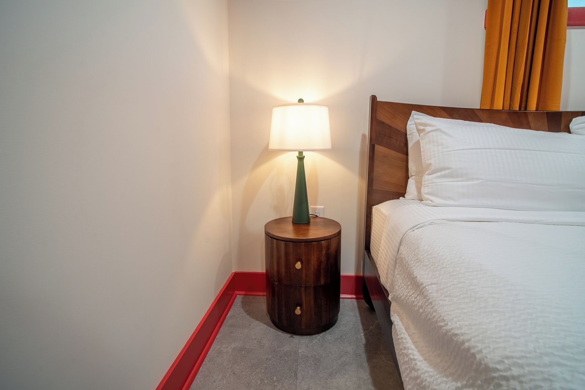 Une chambre d’hôtel avec un lit, une table de chevet avec une lampe et un liseré rouge le long du sol.