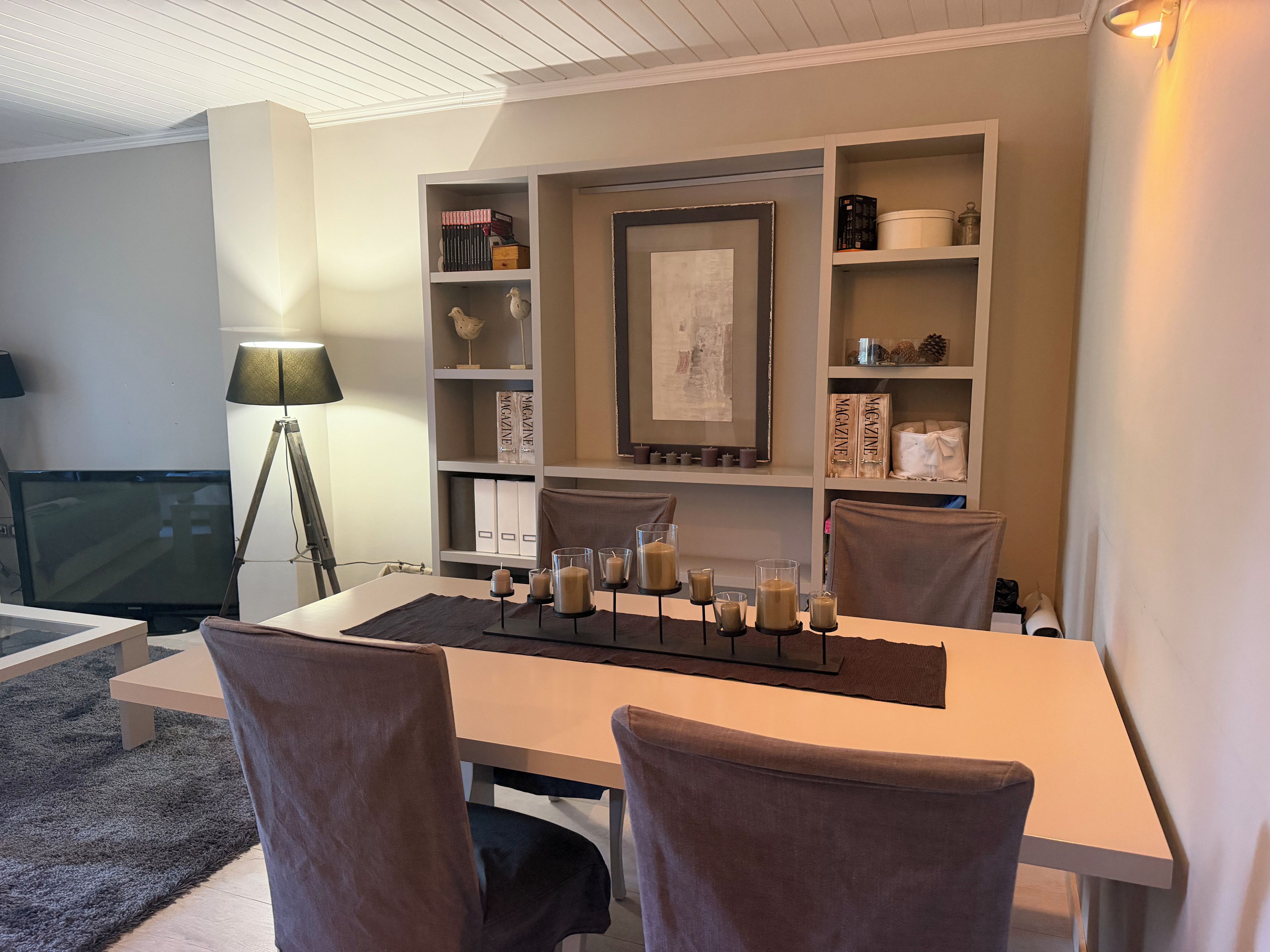 Una sala de estar moderna con una mesa de comedor, sillas, un televisor y una estantería con adornos.