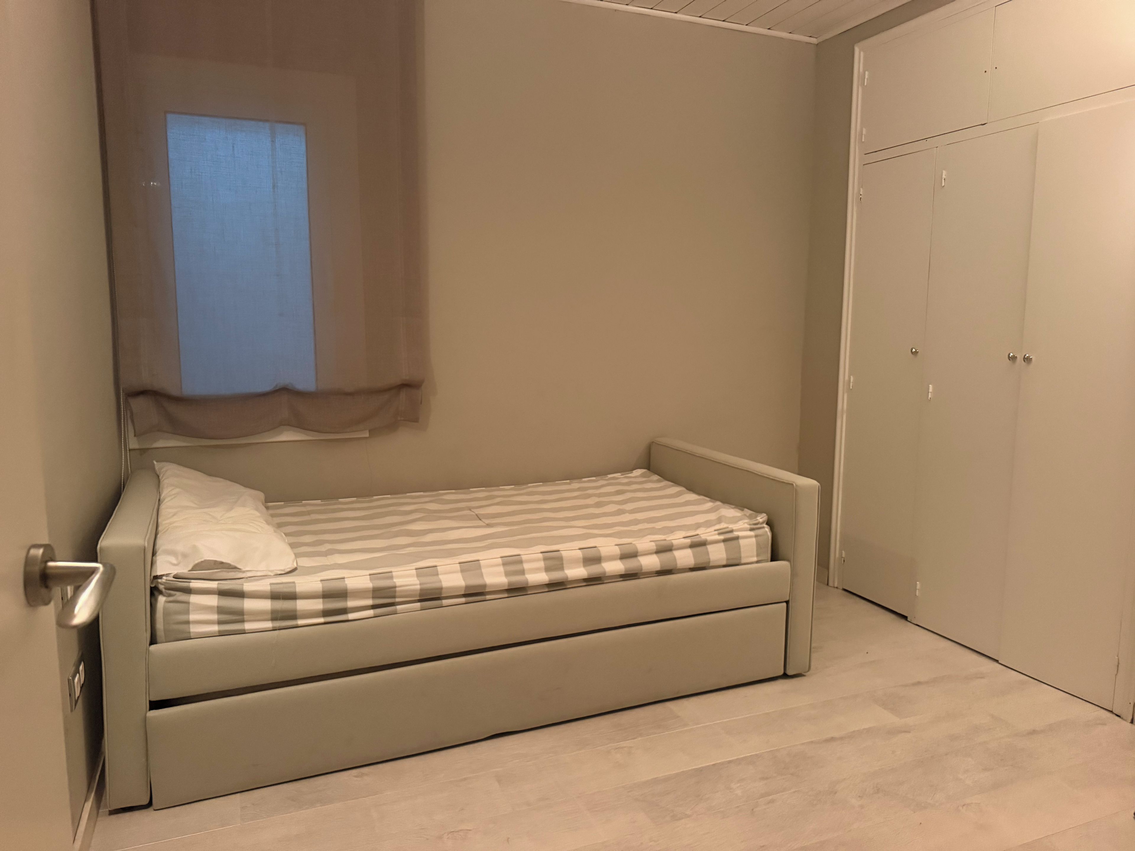 Een kleine eenpersoonskamer met een effen muur, een raam met gordijn en een kledingkast.