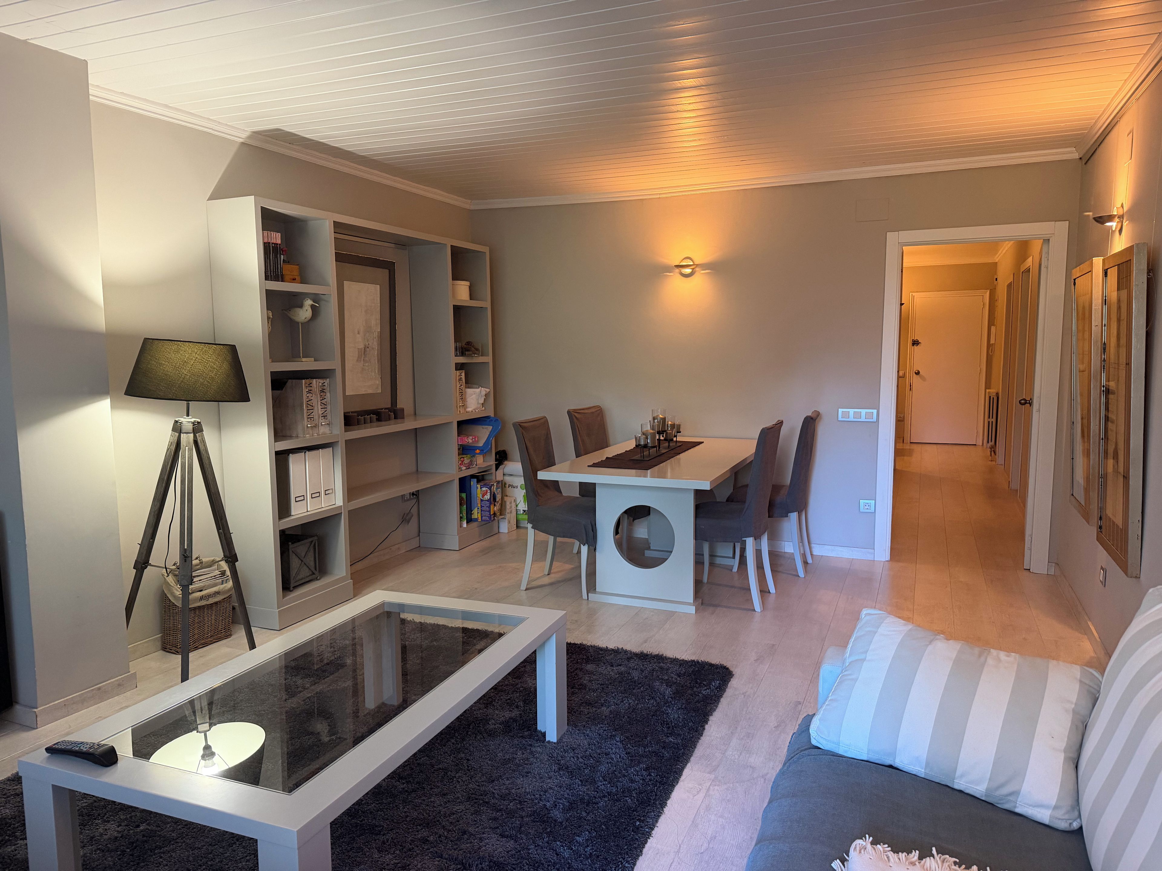 A living room with a glass coffee table, a sofa, a dining table with chairs, a bookshelf, and a lamp.
