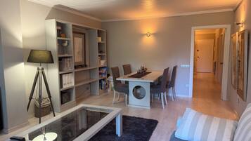 A living room with a glass coffee table, a sofa, a dining table with chairs, a bookshelf, and a lamp.