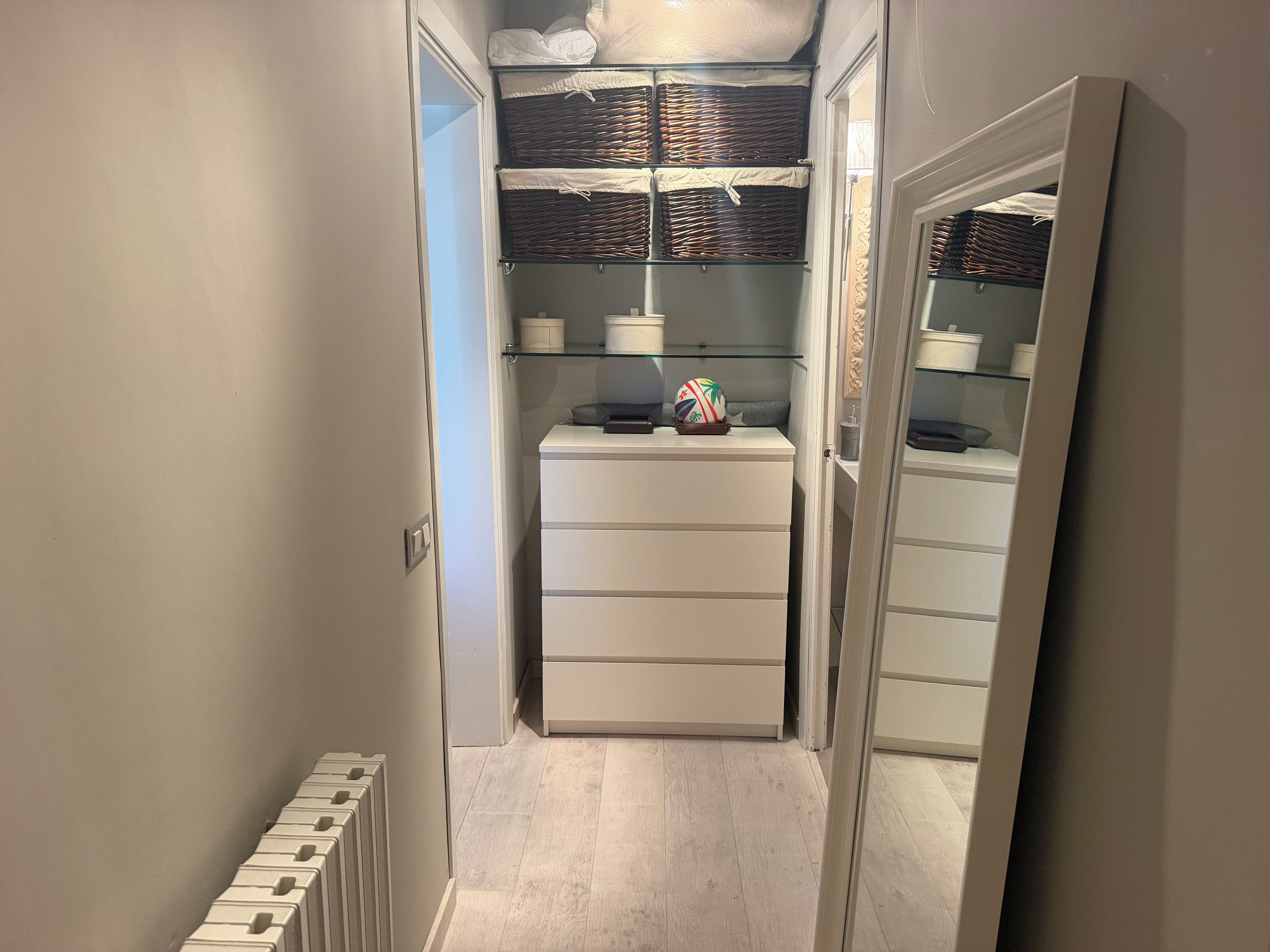 Un pasillo estrecho con un radiador blanco, un mueble blanco con cajones y un estante con cestas y recipientes.