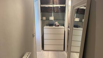 Un pasillo estrecho con un radiador blanco, un mueble blanco con cajones y un estante con cestas y recipientes.