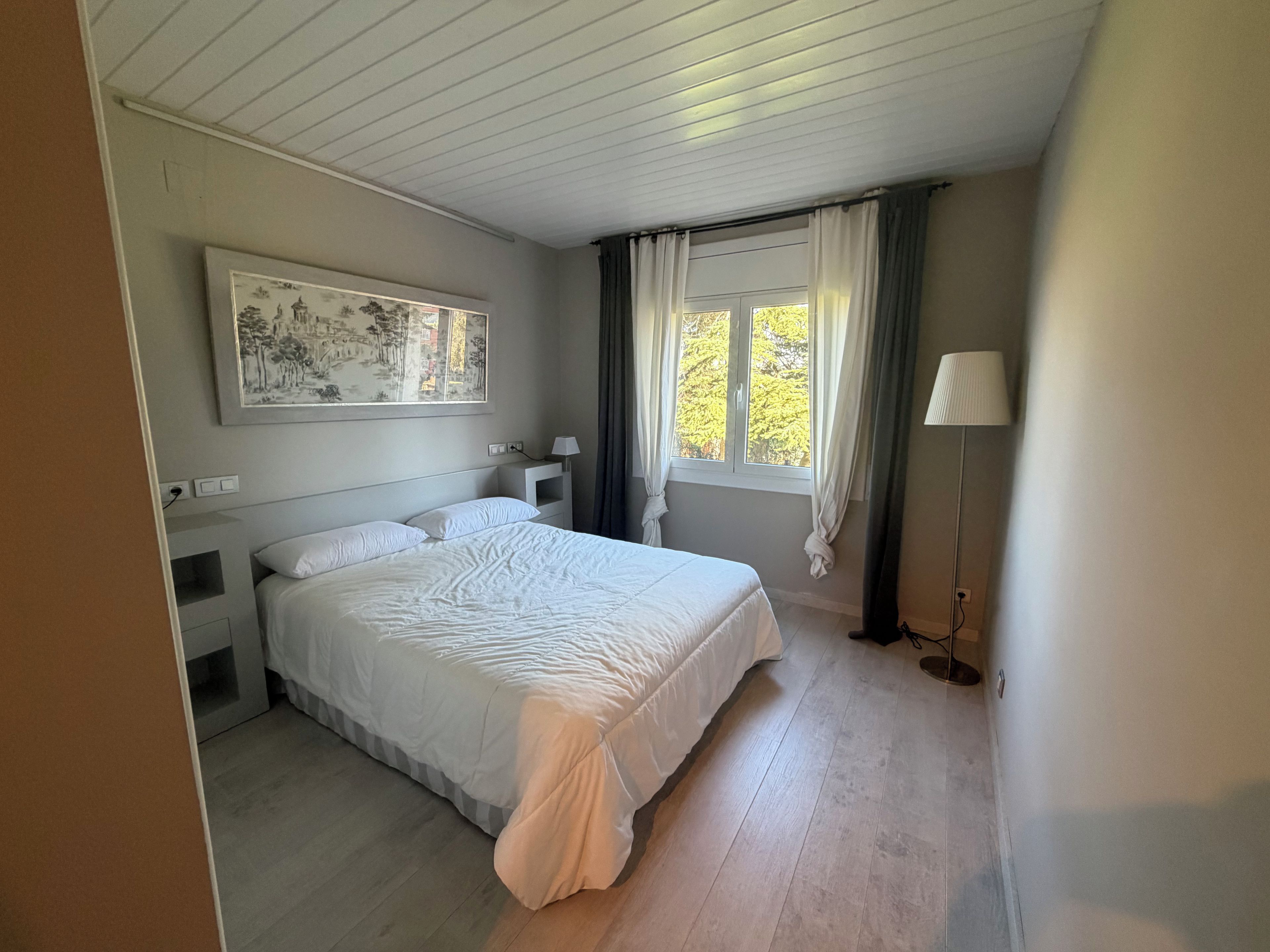 Ein Schlafzimmer mit Bett, Nachttisch, Lampe, einem fenster mit Vorhängen und einem Wandbild.