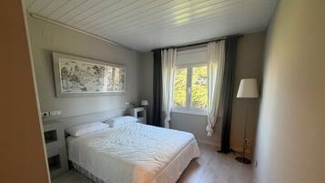 Ein Schlafzimmer mit Bett, Nachttisch, Lampe, einem fenster mit Vorhängen und einem Wandbild.