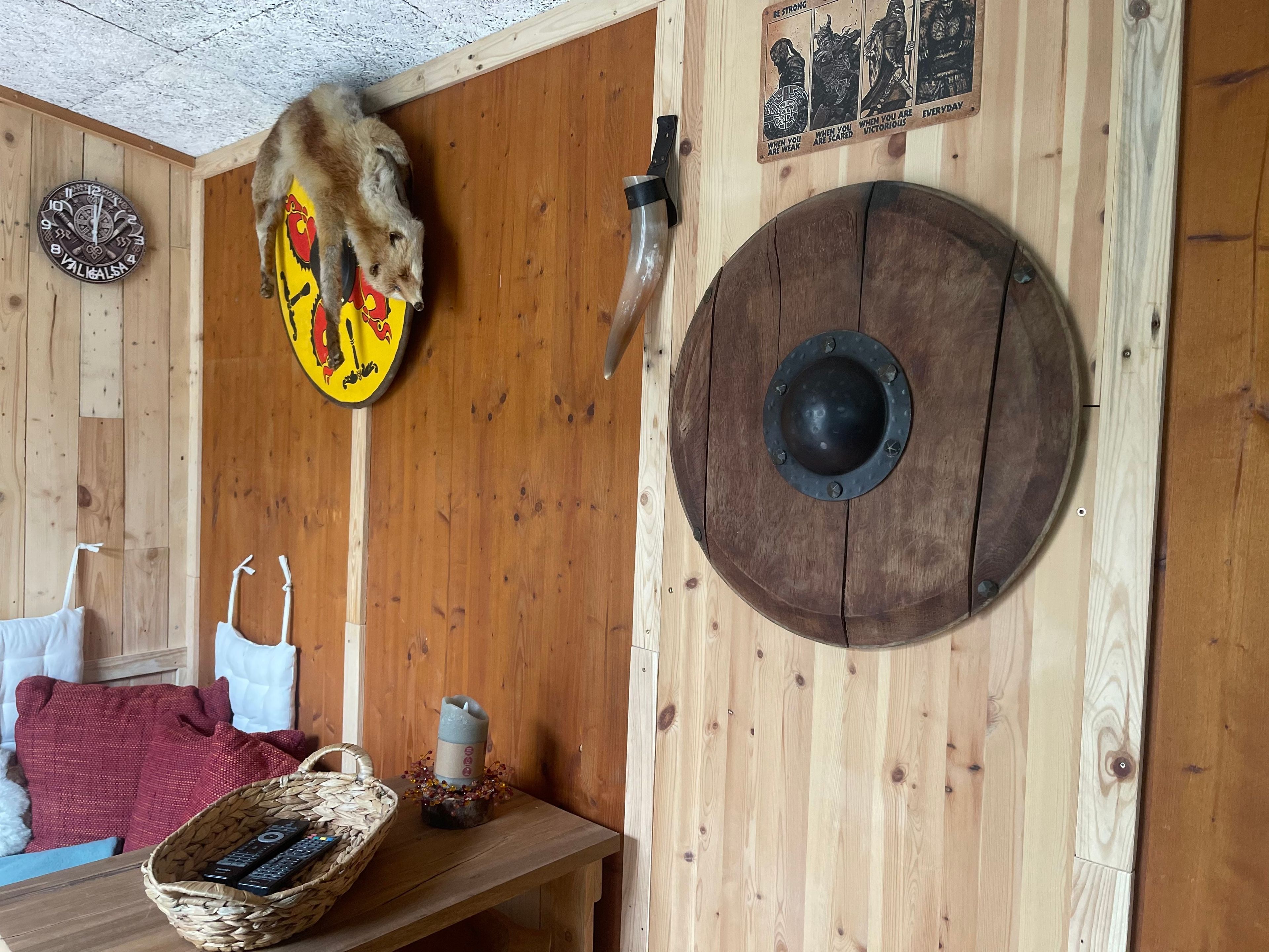 Un intérieur rustique avec un mur en bois, un écu orné d’un motif d’ours, une épée, un bouclier rond, une horloge et un panier contenant des télécommandes.