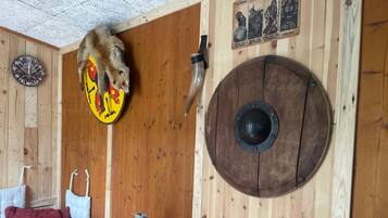 Un intérieur rustique avec un mur en bois, un écu orné d’un motif d’ours, une épée, un bouclier rond, une horloge et un panier contenant des télécommandes.