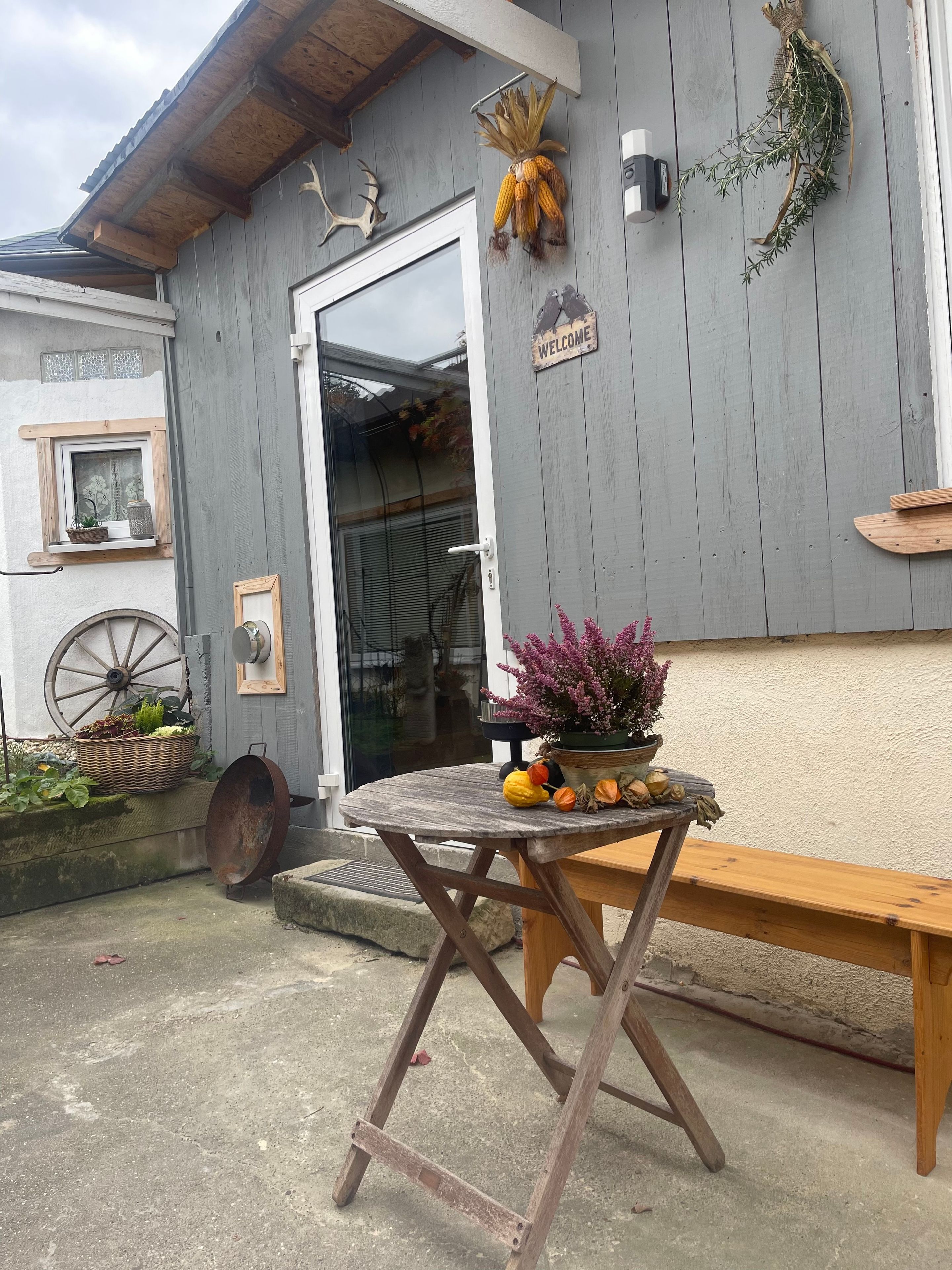 A rustic building with a wooden exterior, a welcome sign, a basket of vegetables, and a table with a flower arrangement.