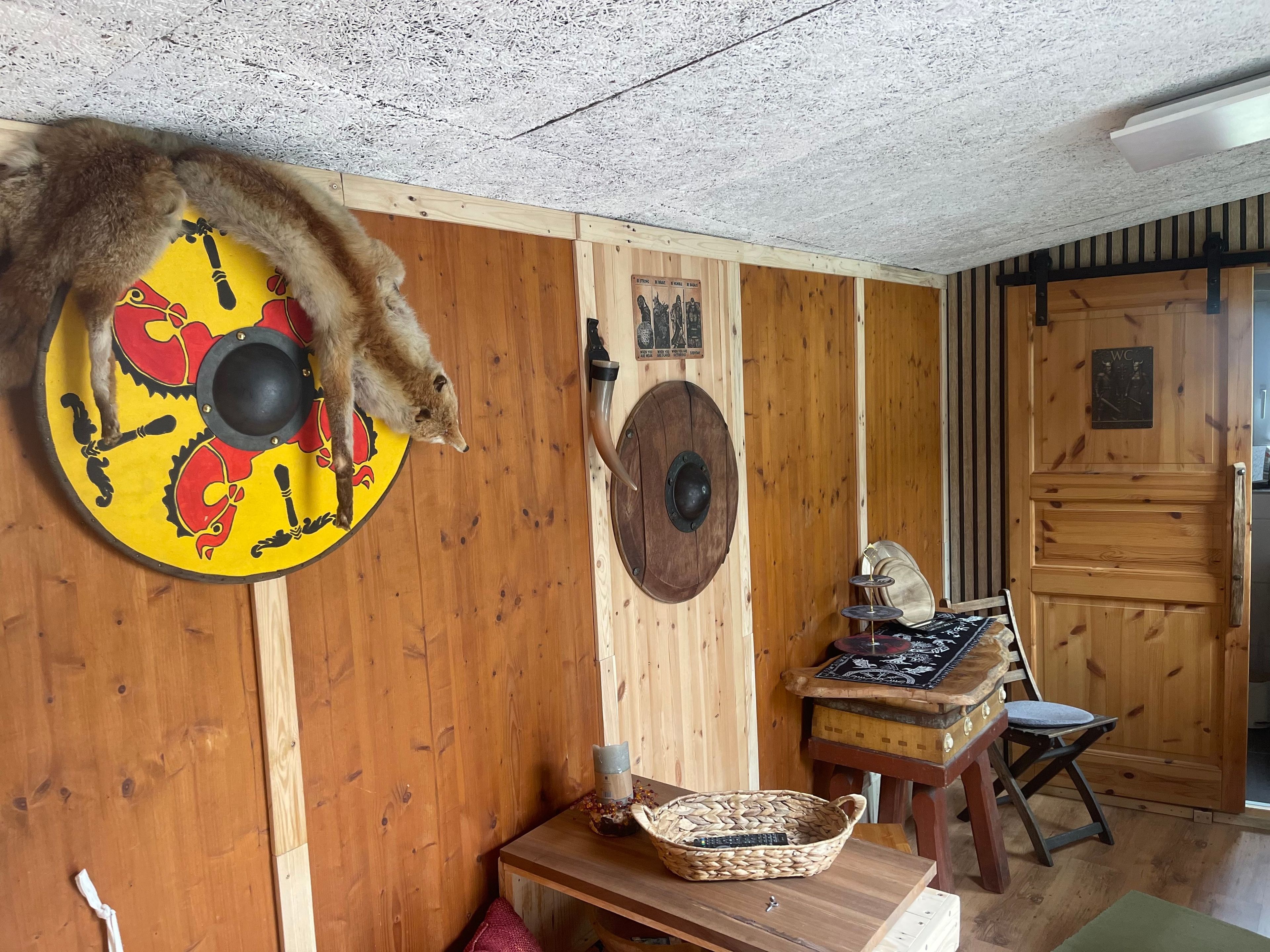 Une pièce aux murs en bois, un bouclier orné d’un motif de crabe rouge, une table en bois avec un panier et une chaise.