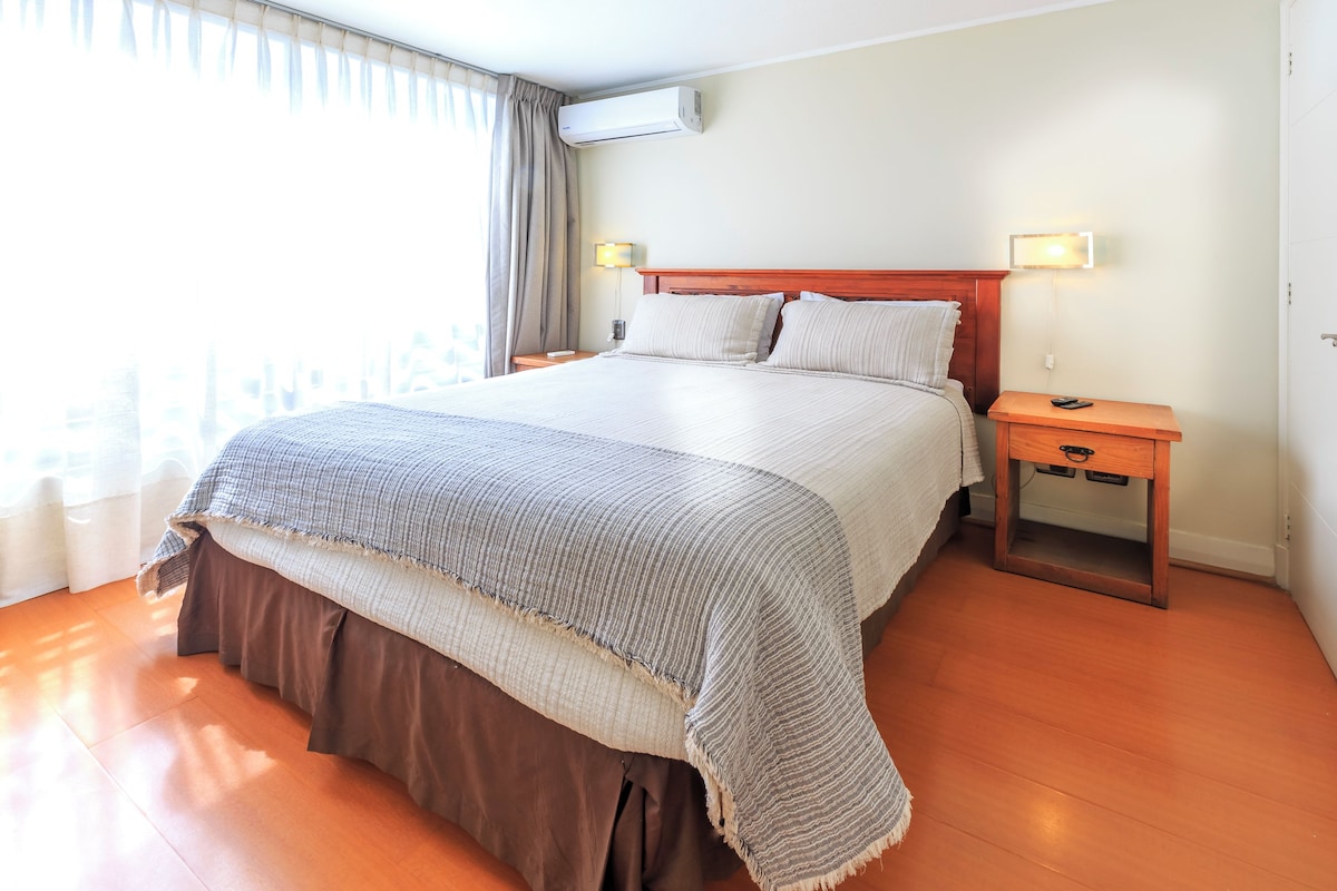 Una habitación de hotel con una cama grande, una mesita de noche y un equipo de aire acondicionado, además de un ventanal con cortinas.