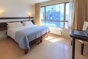 Una habitación de hotel con una cama, un escritorio con una computadora portátil, una ventana con cortinas y un radiador.