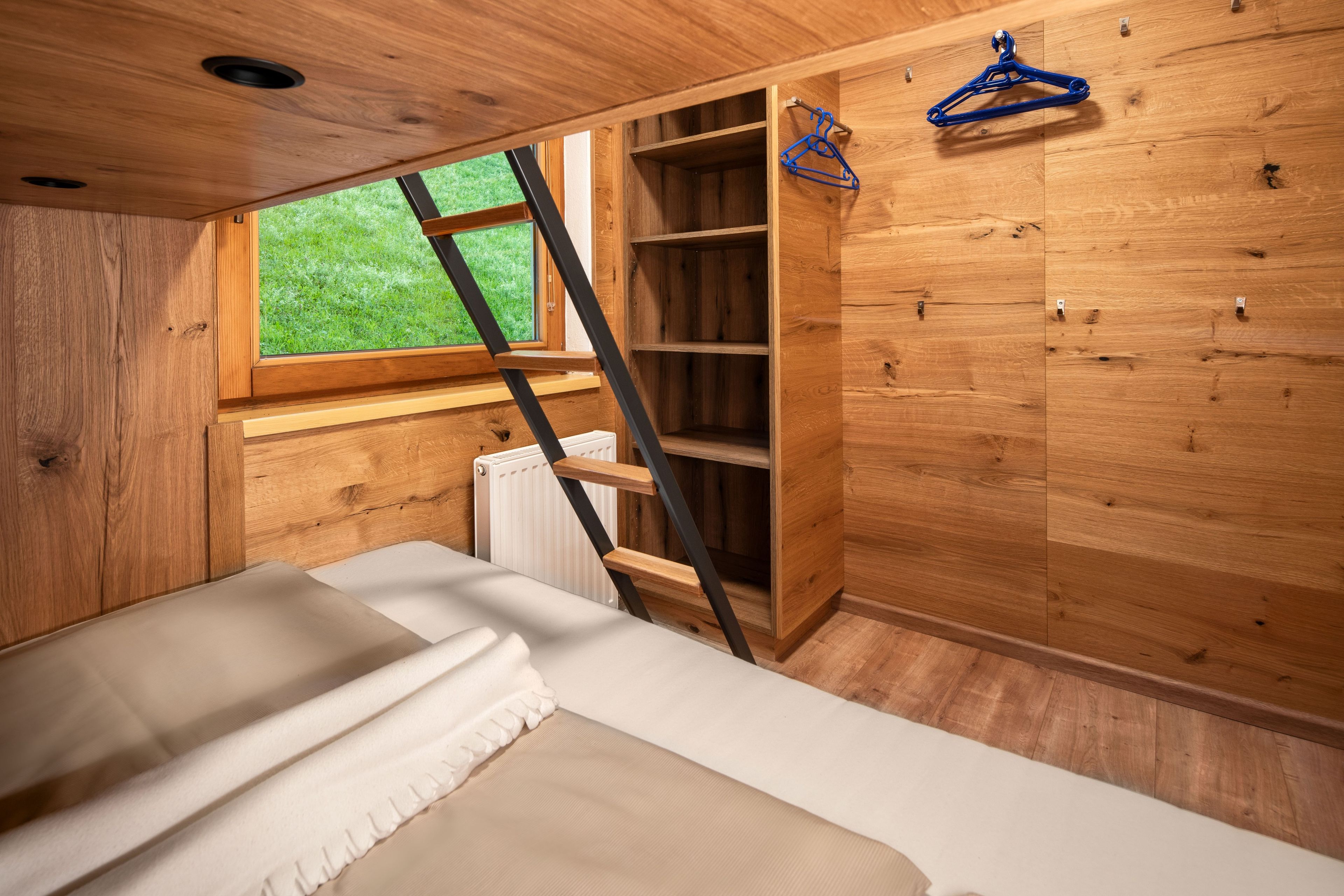 A wooden interior with a bed, a ladder, and a window overlooking greenery.