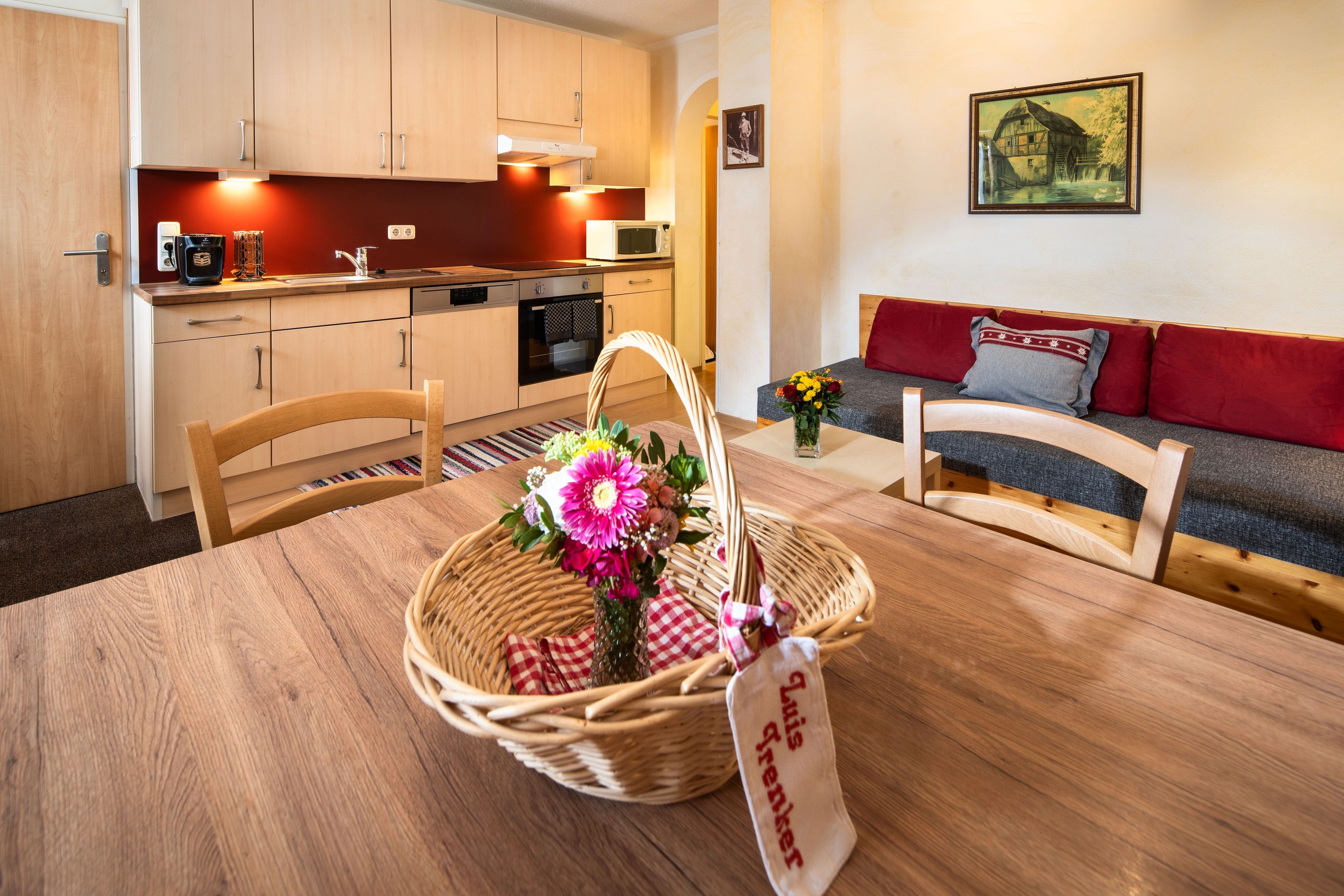 Una habitación acogedora con una mesa de comedor de madera, una cesta de mimbre con flores, un sofá con cojines rojos y una zona de cocina con armarios de madera y un microondas.