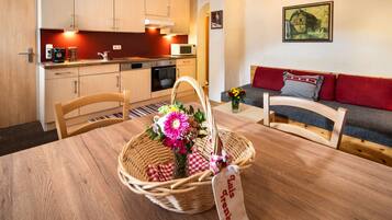 Una habitación acogedora con una mesa de comedor de madera, una cesta de mimbre con flores, un sofá con cojines rojos y una zona de cocina con armarios de madera y un microondas.