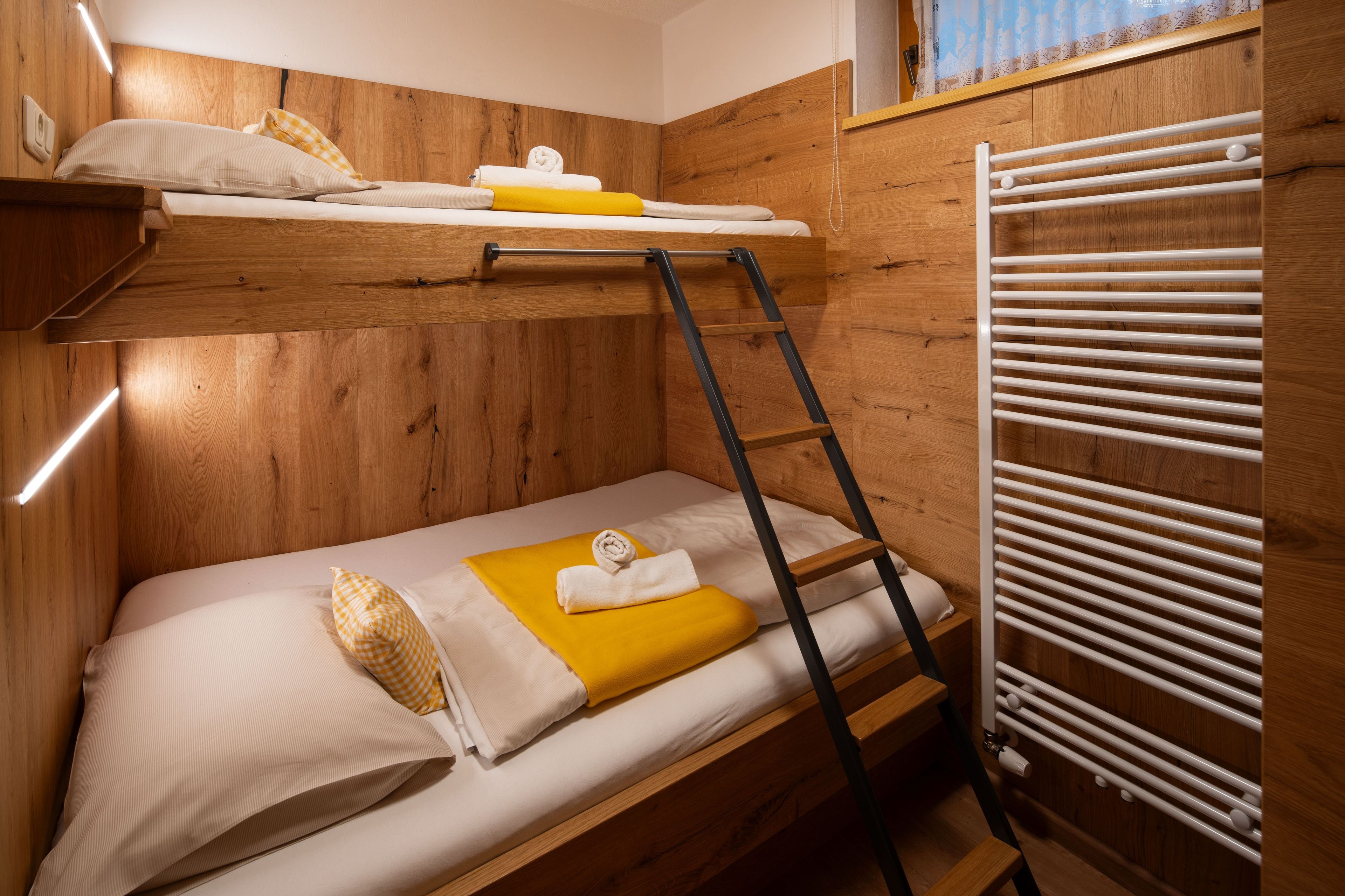 Una cama individual superpuesta con escalera, un radiador blanco y una ventana con cortina.