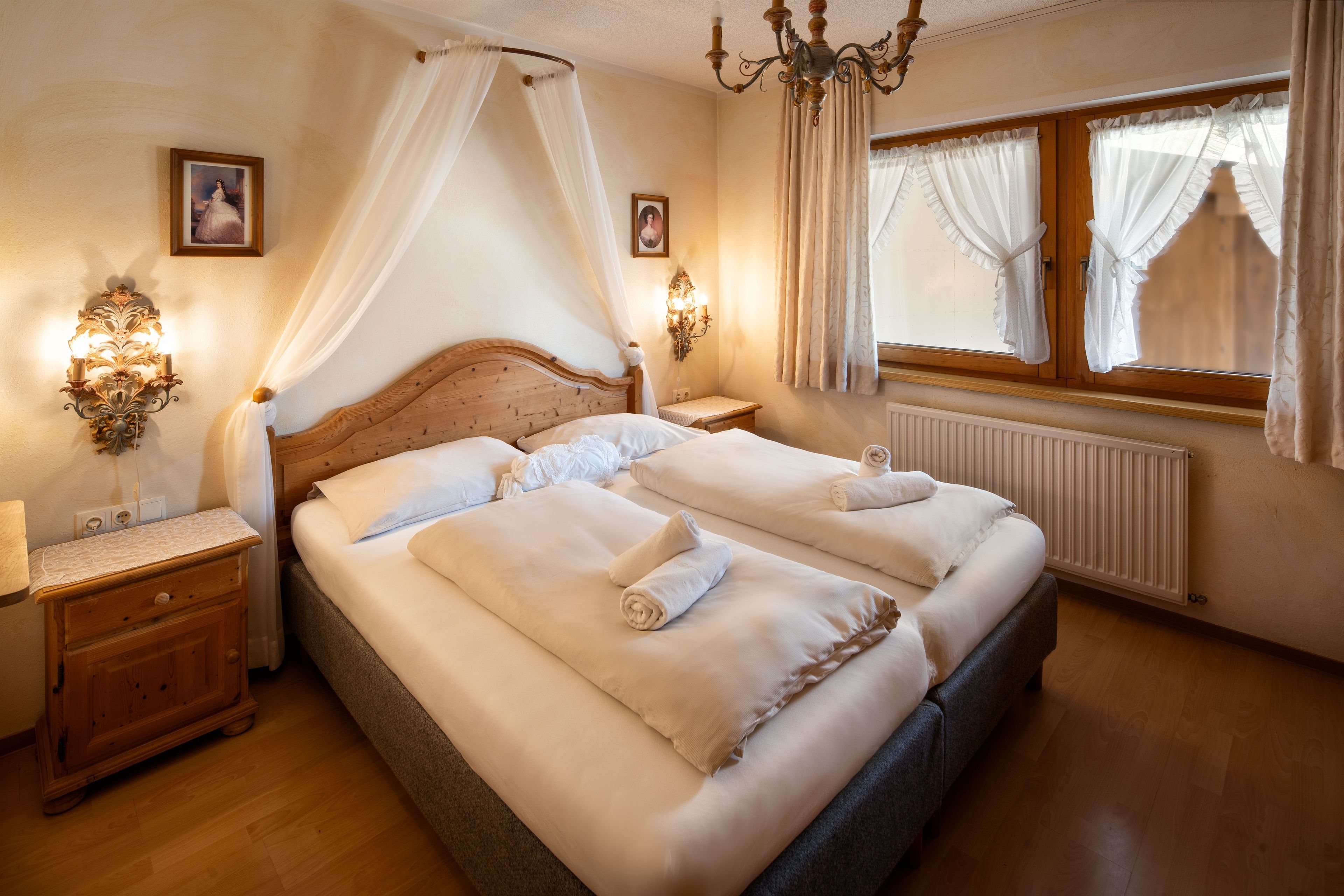 Ein Doppelbett mit weißen Leinen und Handtüchern, ein Holz-Kopfstück, Nachttische mit Lampen, ein Fenster mit Vorhängen und eine Kristallleuchter.