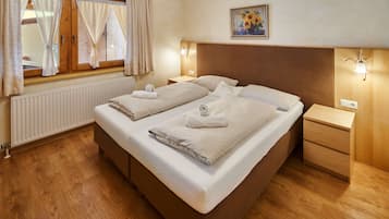 A double bed with two pillows and two towels, a wooden headboard, a bedside table, a nightstand, a radiator, a window with curtains, and a painting on the wall.