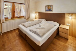 Ein Doppelbett mit zwei Kissen und zwei Handtüchern, ein Holz-Kopfteil, ein Nachttisch, ein Heizkörper, ein Fenster mit Vorhängen und ein Bild an der Wand.