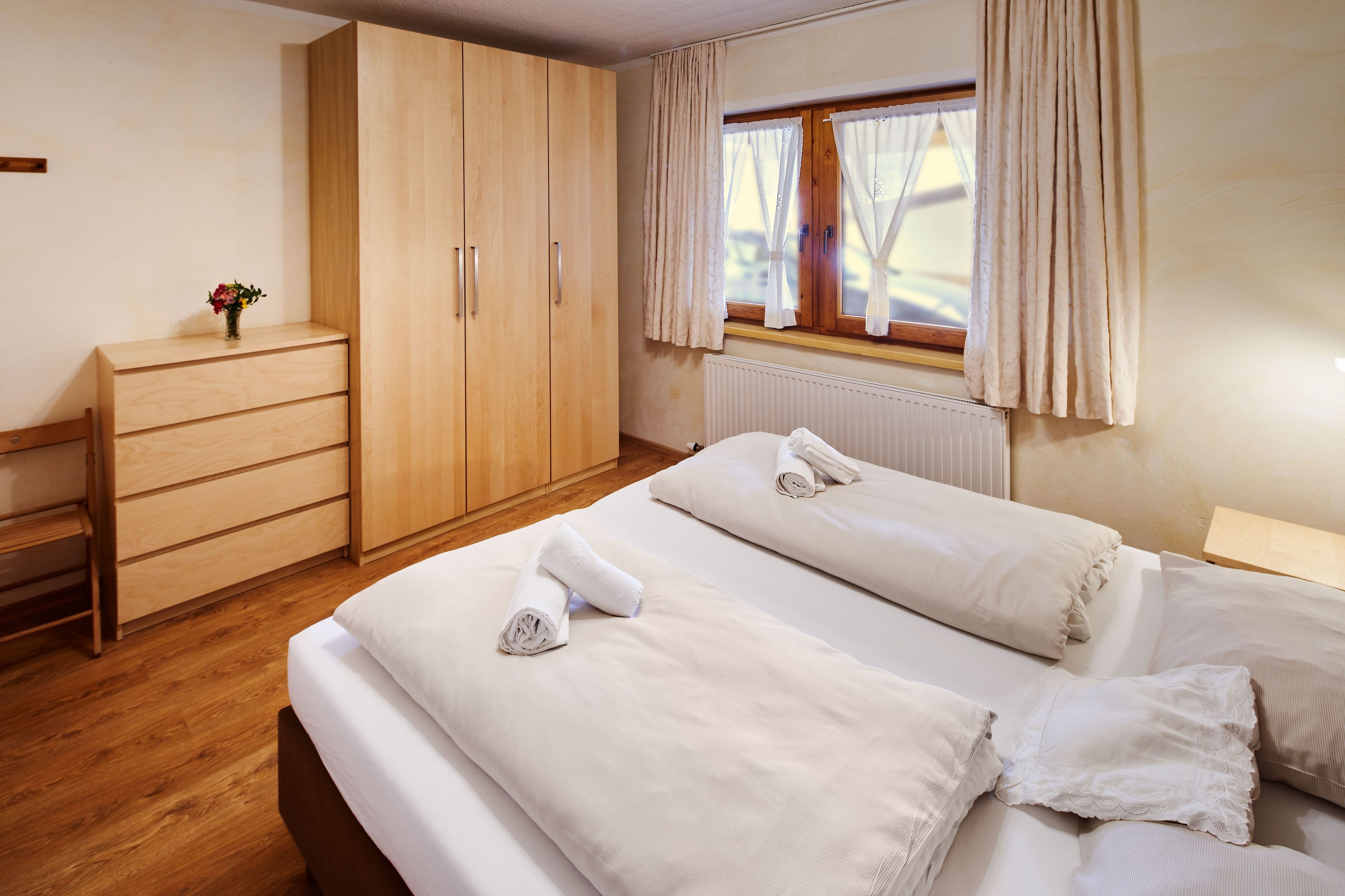 A bedroom with a wooden wardrobe, a bedside table with a vase of flowers, and a window with curtains.