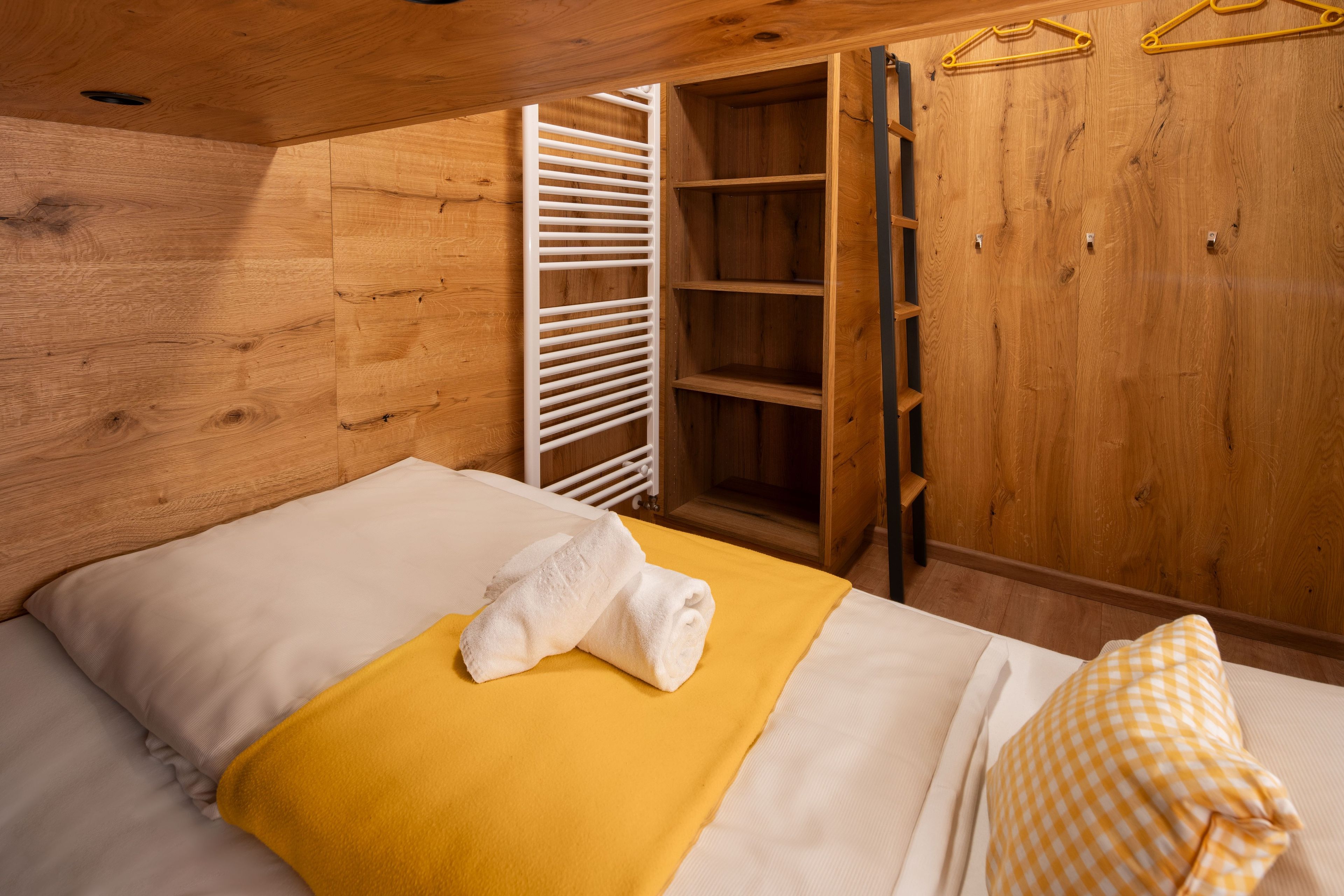 Une pièce en bois avec un lit, une couverture jaune, une serviette blanche, un oreiller à carreaux et une armoire en bois dotée d’une tringle pour suspendre les vêtements.